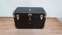 Front view of the black vintage wood footlocker trunk showing metal corner reinforcements and latch locks.