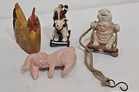 Four assorted figurines shown including wooden chicken, pink ceramic rabbit, wooden rabbit on swing, and Christmas bear on sled.