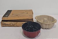 Photo showing vintage Pampered Chef baking set including beige stoneware fluted bundt pan, black/red bar pan, and a boxed item.