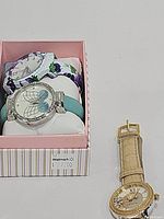 Three watches displayed on white pillows inside a pink box, showing butterfly, floral, and gold-tone watch styles.