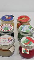 Photo showing all six round glass candles with various Christmas-themed lids including a tree, gift, snowman, gingerbread man, bird, and heart-shaped designs.