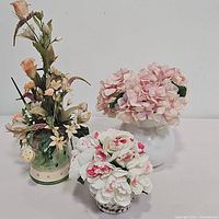 Lot of three floral centerpieces including the watering can, white jar with pink flowers, and white vase with pink and white flowers.