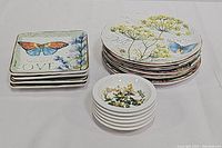 Stack of square butterfly canape plates, round dessert/pie plates with herb garden and butterfly design, and small round botanical bowls on white background
