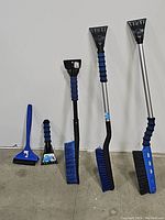 Five snow brushes and ice scrapers arranged side by side on a floor against a white wall, showing different handle lengths and brush colors.