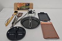 Complete set of grill tools including pans, tongs, spatula, brush, grilling planks, and grill tuner laid out on white surface.
