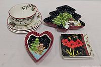 Full set of holiday-themed ceramic pieces including holly plates, bowl, black glazed Christmas tree shaped dishes, and red floral square plates arranged on white surface.