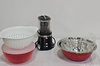 White plastic strainer stacked on red plastic bowl with lid, black brewing tea machine, red and stainless steel nesting bowls with plastic wrap inside