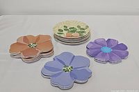 Four sets of melamine plates, three flower-shaped sets in orange, purple, and blue, and one round set with leafy floral design stacked.
