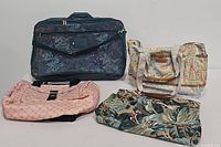 Four overnight bags: blue tapestry rolling bag, pink logo print bag, beige floral cloth bag, large green floral handbag, arranged on floor from front view.