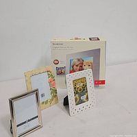 Four picture frames displayed together on white surface: boxed digital frame, white ceramic frame with heart cutouts, decorated rose frame, silver beaded frame.