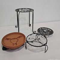Four plant risers displayed including two metal scroll stands, one cast iron flowerpot holder, and one plastic plant caddy.