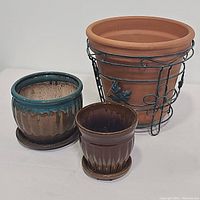 Group photo of three planter pots: large terra cotta pot in metal basket, medium blue-green and brown glazed ceramic pot with saucer, small brown glazed ceramic pot with saucer.