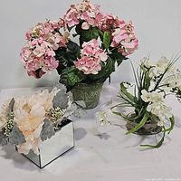 Overall image showing all three floral arrangements together, illustrating the lot's contents including the pink hydrangea in a green pot, white flowers in glass vase, and cream flowers in mirrored container.