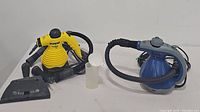 Photo showing both Eureka Enviro Hotshot (yellow) and Scunci ProSteam (blue) handheld steamers with hoses and a small white measuring cup.