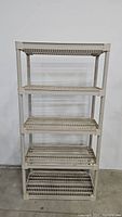 Front view of tan utility shelf with 4 wire mesh shelves against a plain wall