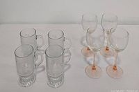 Four clear footed Irish coffee mugs with etched pattern and four pink stemmed goblets shown on white surface.