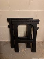 Two black plastic workbench stands folded and leaning against a wall on a concrete floor.