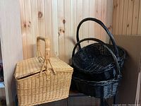 Light tan wicker picnic basket with double hinged lids and central handle, shown with two large black wicker bread baskets with handles nearby.