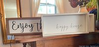 Photo showing two white framed typography signs placed on a wooden surface in front of a window with beige curtains. One sign reads 'Enjoy the', the other 'happy house'.