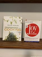 Photo showing two hardcover cookbooks: Professional Cooking by Wayne Gisslen with a white cover and grapes image, and Joy of Cooking with a white cover and large red circle.
