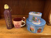 Photo showing all items in the lot on wooden shelf: purple stainless steel water bottle, white Disney princess mug with spoon in handle, and two Christmas-themed cookie tins