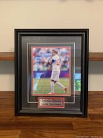 Full view of framed photo depicting Christine Sinclair on a soccer field with a black and red double mat and a gold-edged name plaque beneath.