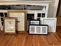 Overview of 9 picture frames in black, white, and gold/bronze colors placed on wooden floor.