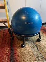 Blue Gaiam stability ball placed on a black rolling stand with caster wheels, on a patterned carpet in a room.