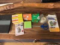 Photo showing various office supplies including staples, tape, scissors, lock, sticky notes, label maker tape, and sheet protectors on a wooden surface.