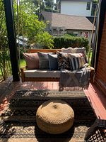 Photo showing 5 outdoor pillows arranged on a outdoor seat, the throw blanket draped on the seat, and the woven pouf in front.
