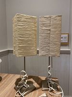 Two IKEA table lamps side by side, showing front view of rectangular cream-white textured paper shades and metallic bases with cords coiled around base.