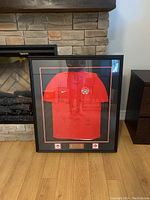 Full view of framed red Team Canada 2020 Olympics women's soccer jersey with signatures, leaning against stone fireplace, wooden floor