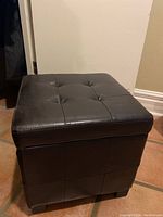 Brown synthetic leather storage ottoman from the side with closed top showing button tufting and grid pattern stitching.