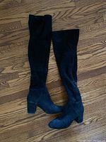 Pair of black suede high boots shown side-by-side on wood flooring to display overall length and shape.
