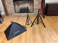Two black tripods and a black Craphy softbox lighting shade set on wood floor in front of stone fireplace.