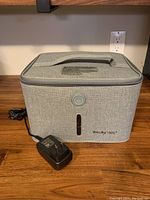 Front view of grey sterilizing bag with power adapter plugged in, showing handle on top and control button on the front.