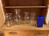 Five vases displayed on a wooden shelf: four clear glass in varying shapes and sizes and one cobalt blue rectangular glass vase.