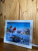 Framed winter scene battery powered light-up wall art showing two cardinals on a snowy branch, a stone bridge, and lit village houses at dusk with a white wooden frame.