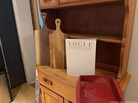 Two wood cutting boards, Vogue Entertaining cookbook, and red silicone bakeware pan on wooden shelf unit.