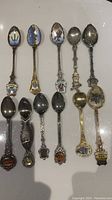 Overview of all 11 collectible souvenir teaspoons laid out on a flat surface, showing bowl designs and handle details in variety of metal finishes including silver and gold tones with enamel and painted decorations.