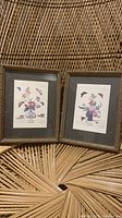 Two vintage botanical prints showing floral arrangements and butterflies, each in dark gray matting and gold ornate frame, placed on wicker chair