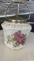 Front side view of the white ceramic biscuit jar with metal lid and handle, showing floral rose motif and embossed pattern.