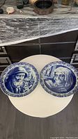 Photo of the two blue and white vintage Royal Delft Boch wall plates on a round white table showing each plate's portrait.