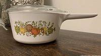 Side view of white CorningWare saucepan with 'Spice of Life' vegetable pattern and long handle on a wooden surface