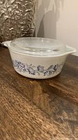 A full side view of the white Pyrex casserole dish showing the blue Colonial Mist flower pattern and clear glass lid on a wooden surface.