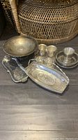 Seven vintage silver plated items on floor with wicker furniture background, including trays, cups, and bowls showing their shapes and patina.