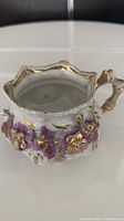 A small white porcelain dish with intricate raised floral decorations in purple and gold. Shows detailed close-up of one side with gold accented ruffled edge and handle.