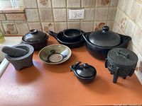 Photo showing an assortment of international kitchen items on a kitchen counter: granite mortar and pestle, black ceramic pots with lids, decorative ceramic bowl, cast iron vessel.