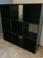 Photo showing black cube storage shelving unit with nine square compartments in a 3x3 layout, constructed of black finished wood or composite material, placed against a wall on wooden floor.