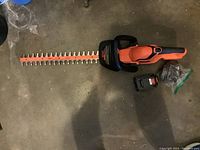 Orange and black Black & Decker cordless hedge trimmer with long blade, front handle, battery and charger laid out on concrete floor.